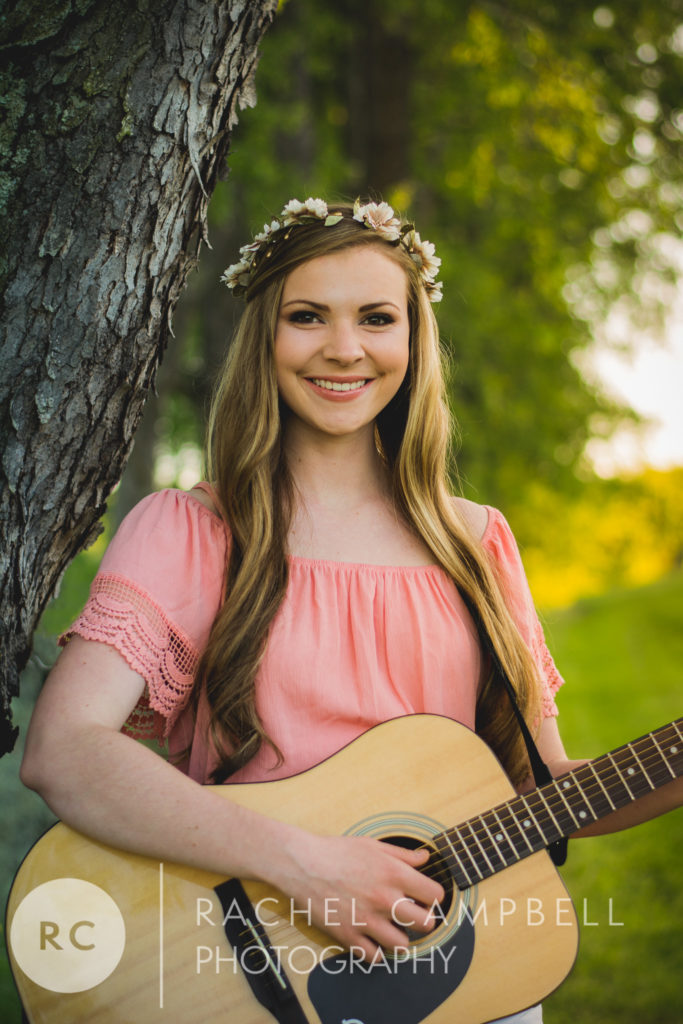Senior portrait of a young woman playing guitar while leaning against a tree at sunset in Solon Ohio