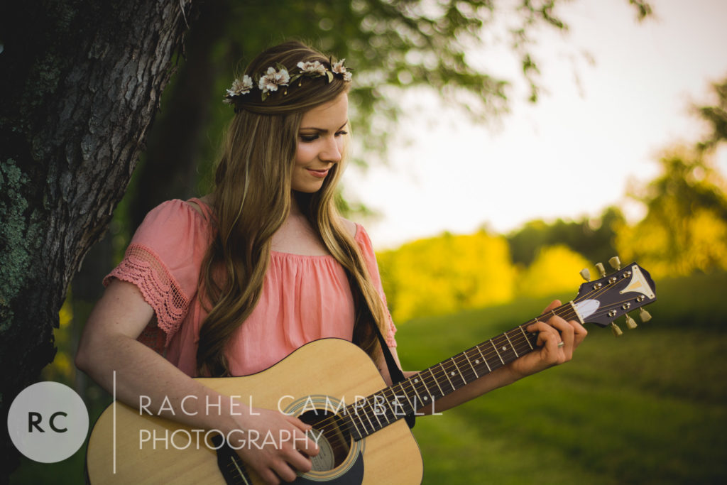 Senior portrait of a young woman playing guitar while leaning against a tree at sunset in Solon Ohio