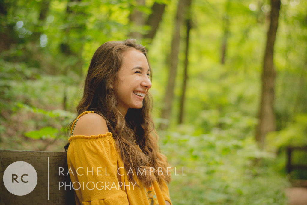 senior portrait of a young woman sitting on a bench laughing in the woods in Solon Ohio.
